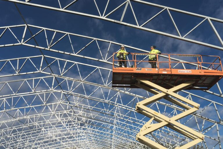 Two workers on a scissor lift at a construction site, framed by a metal structure against a blue sky.