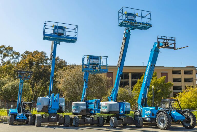 Teleskoparbeitsbühne mieten – Outdoor scene with multiple blue aerial lifts and heavy machinery in a sunny industrial ar